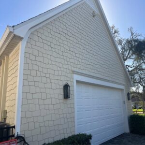 coastal style house with light tan shingle siding and white garage door well maintained landscaping and clear blue sky