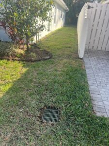 drainage grate on a grassy area beside a house with a fence and paver walkway