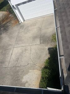 concrete driveway with visible cracks and weeds along the edge and gutter system positioned on the roofline