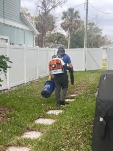 a worker using a leaf blower and carrying a collection bag while walking through a yard with stepping stones