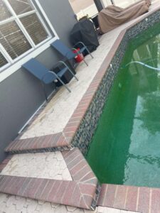 green pool water with blue vacuum hose alongside brick and stone patio and two chairs near a barbecue grill