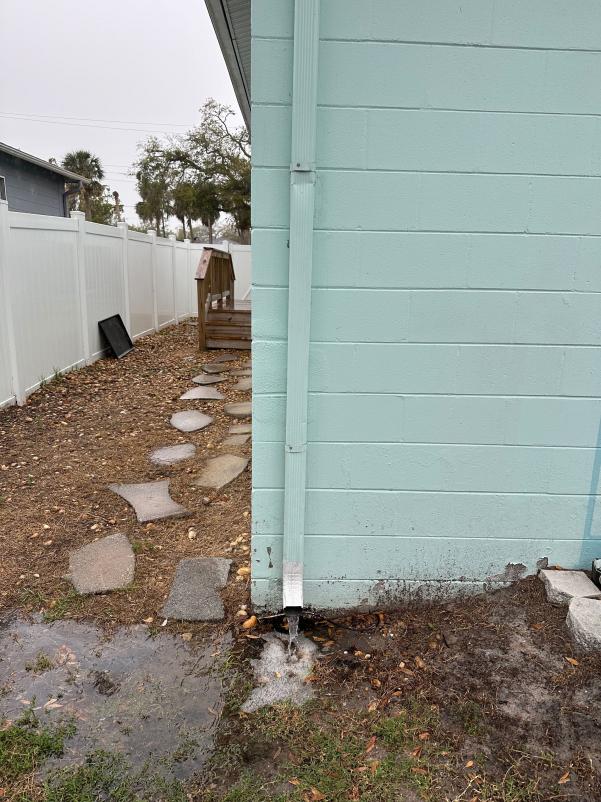 gutter downspout draining water onto the ground with some mud and debris accumulating around the base of the wall