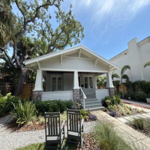 a charming white house with a porch surrounded by lush landscaping and two green chairs on the lawn