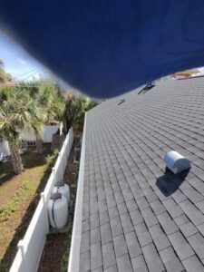 cleaning a dark asphalt roof with visible vents and white fence surrounding a landscaped area with palm trees