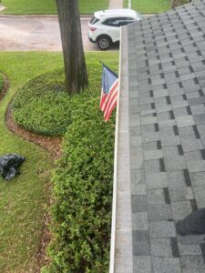 gutter cleaning being performed on a residential roof with an american flag visible and landscaping in the background