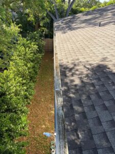 gutter cleaning along the edge of a roof showing debris buildup and vegetation along the side of the house