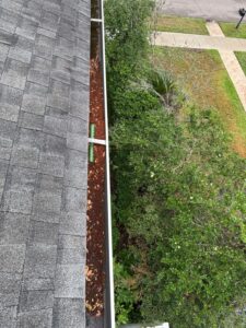 clogged gutter with leaves and debris near a roof edge surrounded by greenery and a clear sidewalk below
