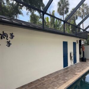 cleaning a white wall and blue doors near a pool area with pavers and foliage in the background