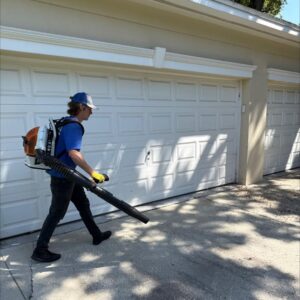 man using a backpack blower to clear debris from a driveway in front of a garage door