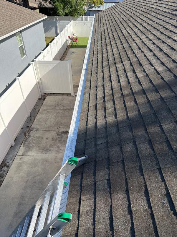 gutter cleaning in progress using a ladder on a sloped roof with concrete and grassy area visible below
