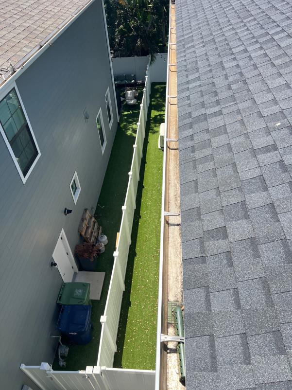 gutter cleaning along roof edge with a view of a narrow green yard and fence on one side