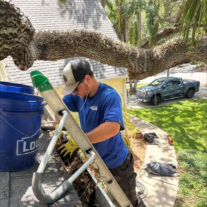 man clearing debris from gutters while using a ladder and wearing gloves with a blue bucket nearby