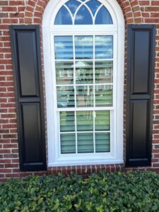 clean window with black shutters on a brick wall surrounded by greenery and clear reflection of the sky and house