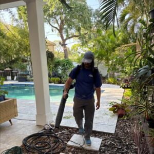 man using a leaf blower to clear debris near a swimming pool surrounded by tropical plants and landscaping