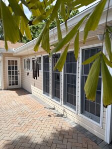 house with multiple windows and a brick patio featuring a door and wind chimes surrounded by greenery