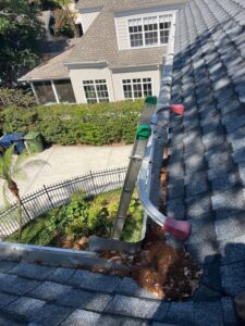 cleaning gutters using a ladder and tools with debris accumulation visible near the plants and landscaping below
