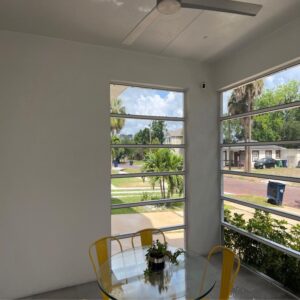 a bright indoor space with large windows a glass table and yellow chairs overlooking a garden and street view