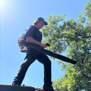 a worker using a leaf blower on a roof removing debris under a clear blue sky with trees in the background