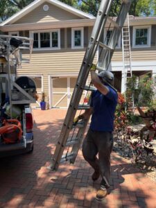 worker carrying a ladder near a truck parked in a driveway surrounded by landscaping and a house in the background