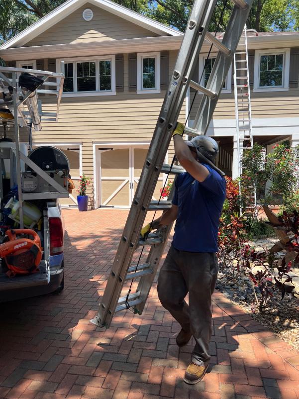 worker carrying a ladder near a truck parked in a driveway surrounded by landscaping and a house in the background