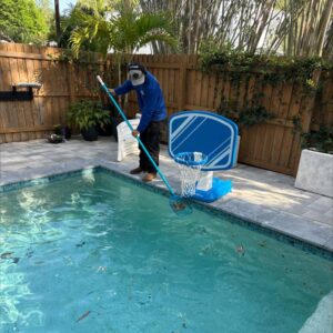 man cleaning pool with a net removing debris from the water surrounded by a wooden fence and tropical plants
