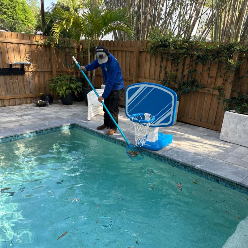 man cleaning pool with a net removing debris from the water surrounded by a wooden fence and tropical plants