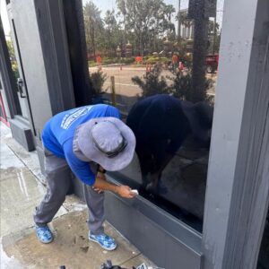 a worker cleaning a large glass window using a squeegee while wearing a wide-brimmed hat and blue shirt