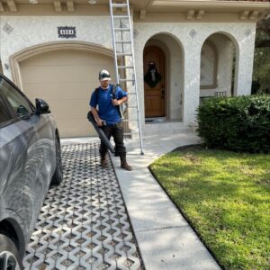 worker using a leaf blower to clear debris from a pathway with a ladder positioned nearby and a car in the driveway