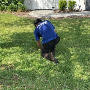 a worker in a blue shirt kneeling on grass possibly inspecting or cleaning an area near a house
