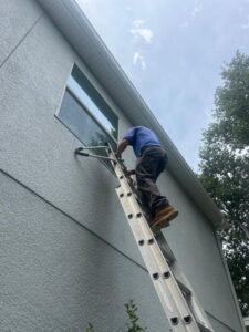 man on ladder cleaning a large window with a brush near a house wall and trees in the background