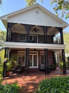 two-story house with a porch featuring decorative iron railings and a brick patio surrounded by greenery and potted plants