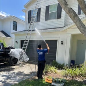 worker pressure washing the exterior of a house using a ladder and hose while a truck is parked nearby