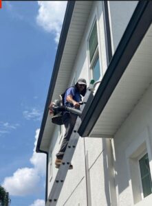 a worker using a ladder to clean gutters with a vacuum attachment on a sunny day