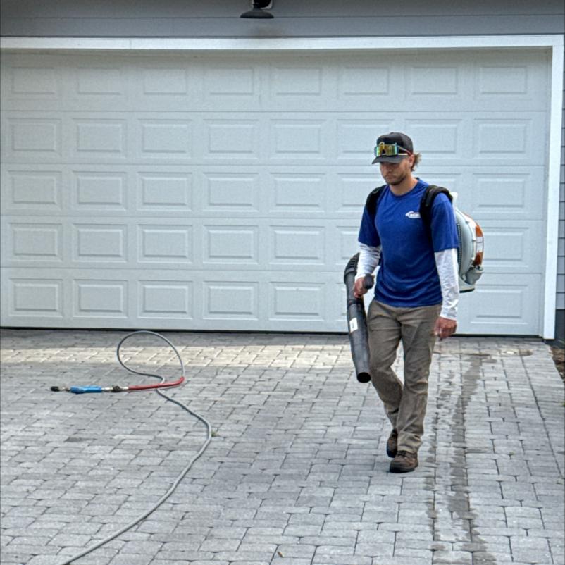man carrying a leaf blower walking on a paver driveway with hoses lying on the ground near a garage door