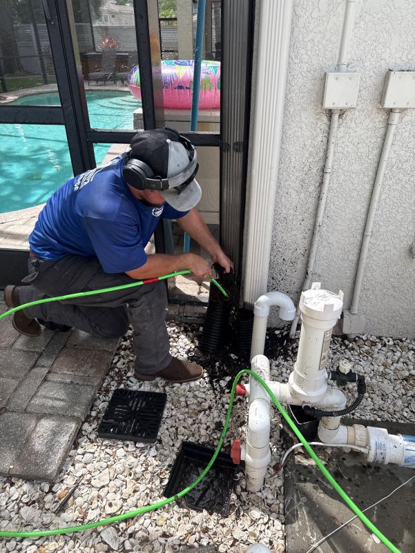 employee cleaning a drain using a hose and equipment near a pool area with decorative stones around the pipes