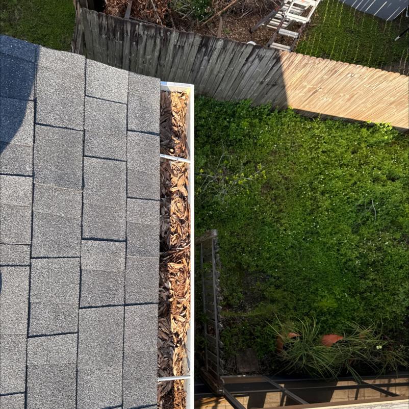 gutter cleaning service clearing debris from a gutter filled with leaves near a roof edge and green backyard visible