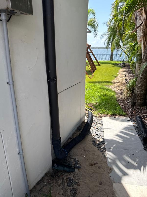 drainage system installation with black hoses extending to landscaping near a waterfront location surrounded by grass and palm trees
