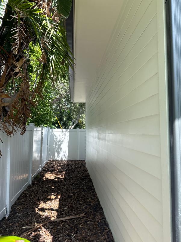 cleaning a side of a house with light colored siding surrounded by a fence and overgrown plants