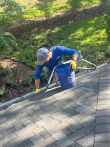 gutter cleaning being performed by a worker using a bucket and ladder on a roof with shingles