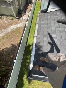 cleaning gutters from a rooftop while wearing work boots and standing on a gray shingle roof
