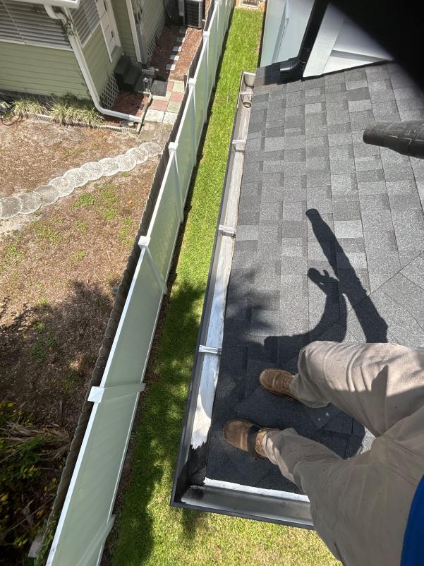 cleaning gutters from a rooftop while wearing work boots and standing on a gray shingle roof