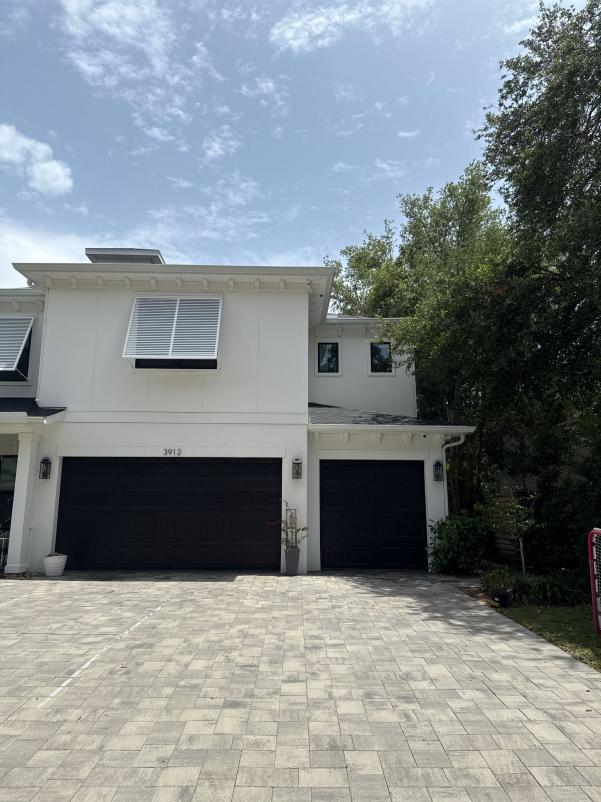 two garage doors with a stone paved driveway and a house with modern architecture surrounded by greenery and a clear sky
