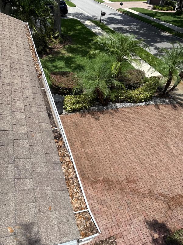 gutter cleaning showing leaves and debris along the edge of a roof with a brick driveway and landscaping visible