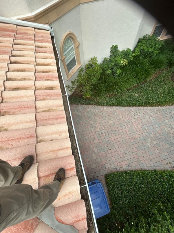 worker on a tile roof preparing to clean gutters with a view of landscaped area and paver driveway below
