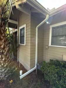rain gutter downspout installation on a house with tan siding surrounded by greenery and palm trees