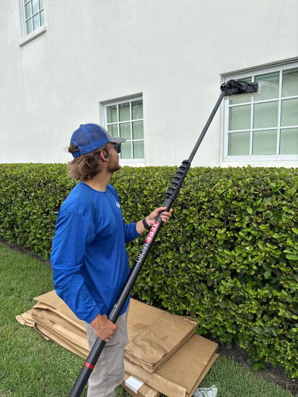 window cleaning using a long pole with a brush on a white building near trimmed hedges and cardboard boxes on the ground