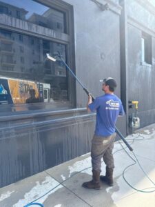 man cleaning windows with a long pole water flowing down wall and equipment placed nearby on the ground