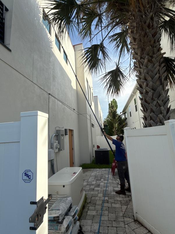 someone using a long pole for cleaning the side of a building near palm trees and a white fence