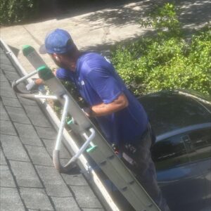 worker cleaning gutters from a ladder using tools on a roof with surrounding foliage and a parked car below