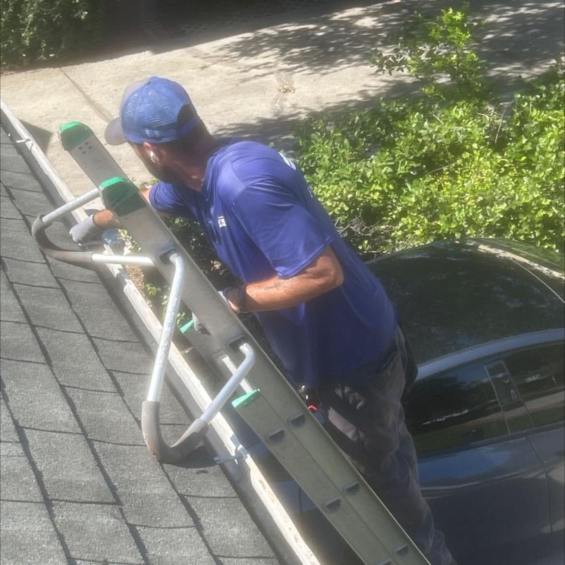 worker cleaning gutters from a ladder using tools on a roof with surrounding foliage and a parked car below